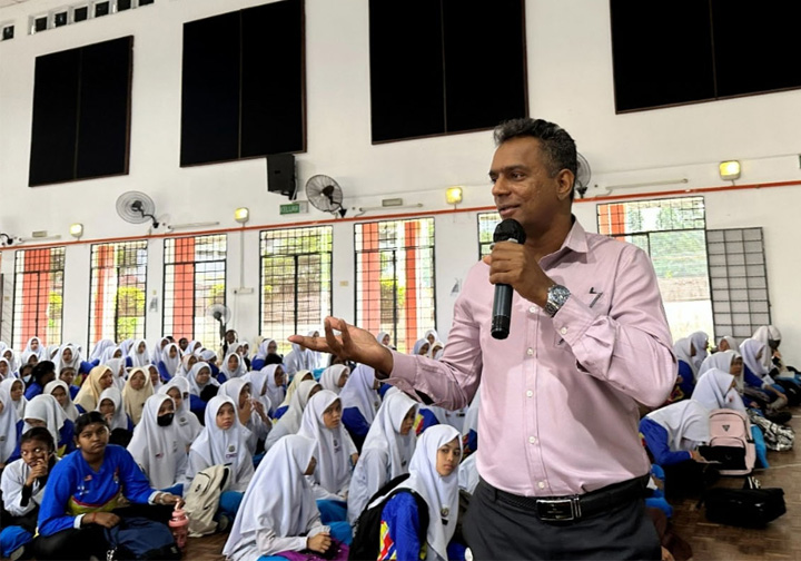 An Intertek colleague holding a microphone addresses a hall full of school students. 