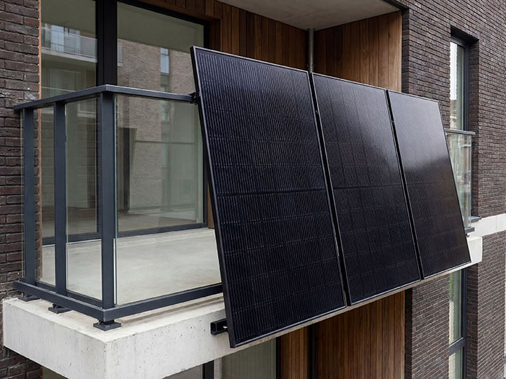 Three black PV solar panels mounted on a balcony with glass railings of a modern apartment building