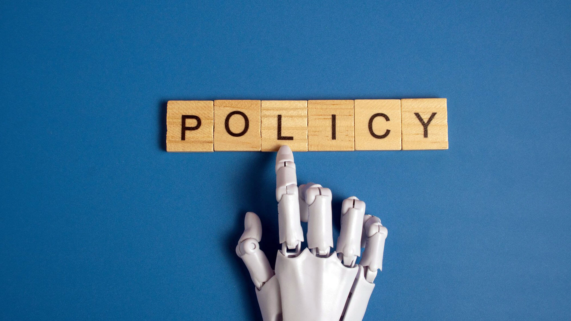 White robotic hand pointing to the word policy on wooden blocks over a blue background representing artificial intelligence ethics and government regulations. 